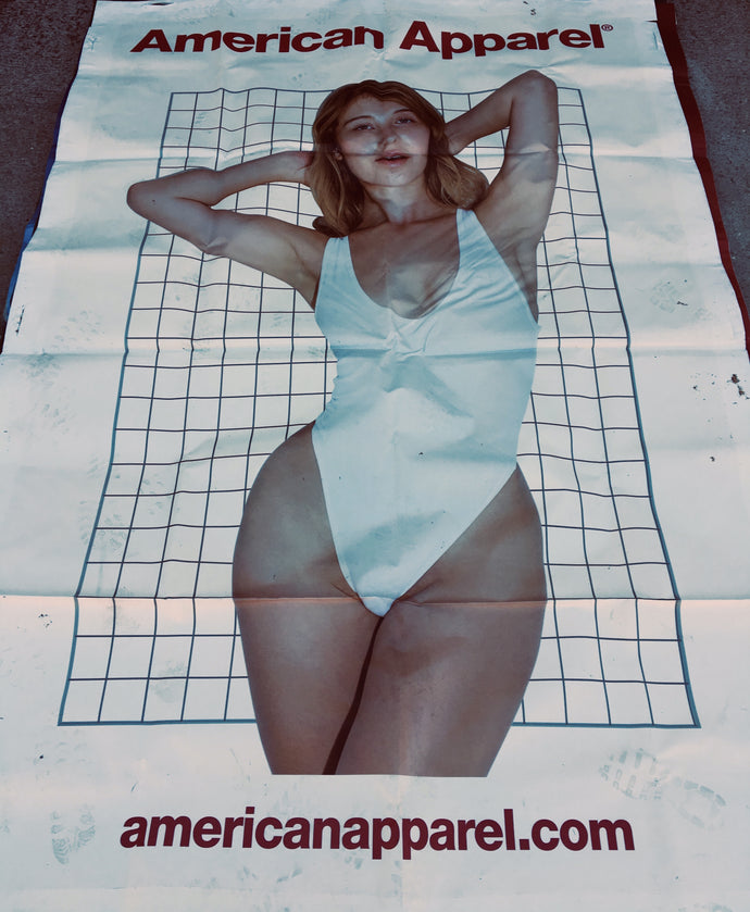 An American Apparel billboard with a girl in a white swimsuit standing against a wire grid background. 